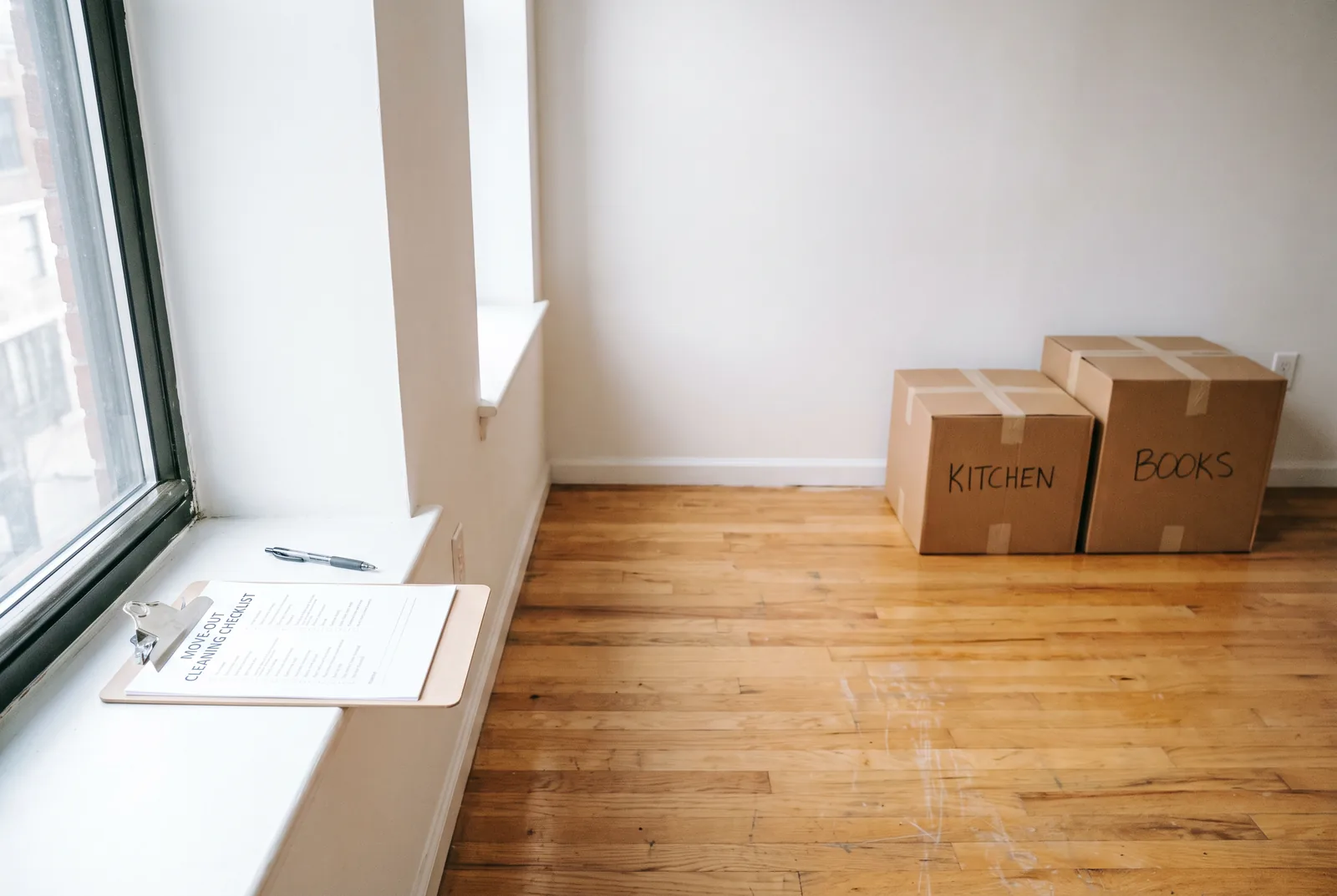 A printed move-out cleaning checklist on a clipboard resting on a windowsill in an empty room with labeled moving boxes